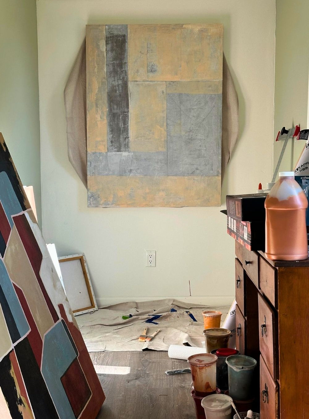 Art studio with artwork on easels, a painting on the wall, and various art supplies on a floor covered with a drop cloth.